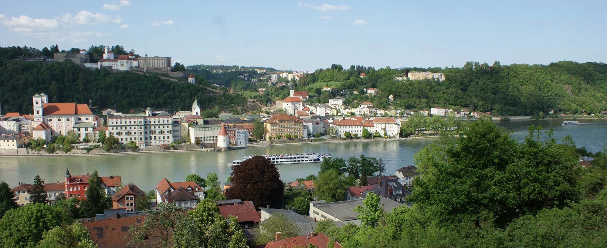 Blick über Passau mit Altstadt, Fluss und Ausflugsschiff, umgeben von bewaldeten Hügeln