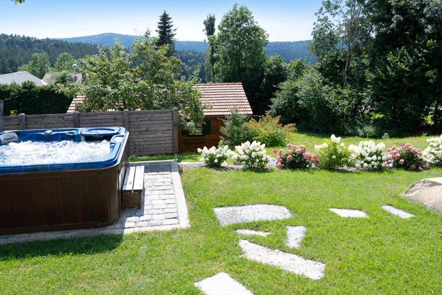 Sprudelnder Außen-Whirlpool auf der Rasenfläche mit blühenden Sträuchern und Blick auf bewaldete Hügel