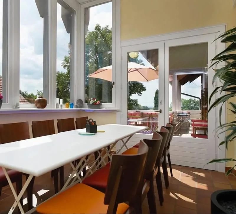 Wintergarten mit langer Tafel, farbigen Stühlen und Tür zur Terrasse