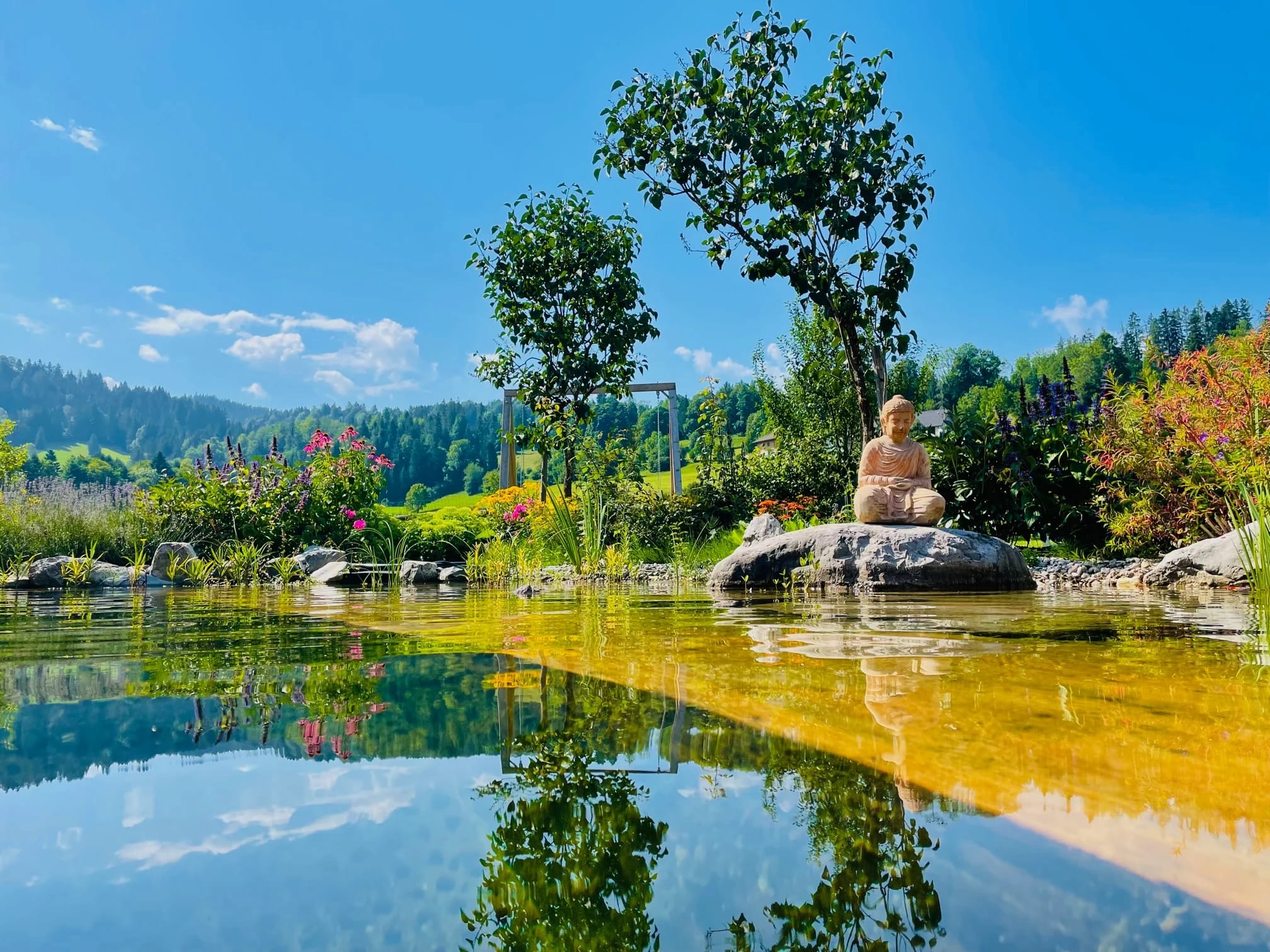 Naturbadeteich mit Buddha-Figur auf einem Stein und blühenden Ufern