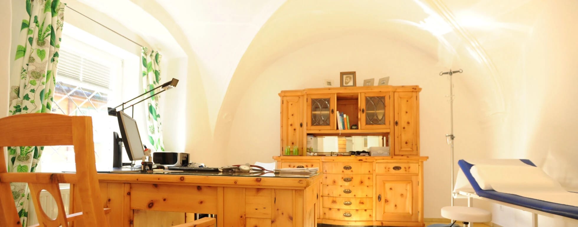 Behandlungsraum mit Gewölbedecke, Möbeln aus hellem Holz und Behandlungsliege am Fenster