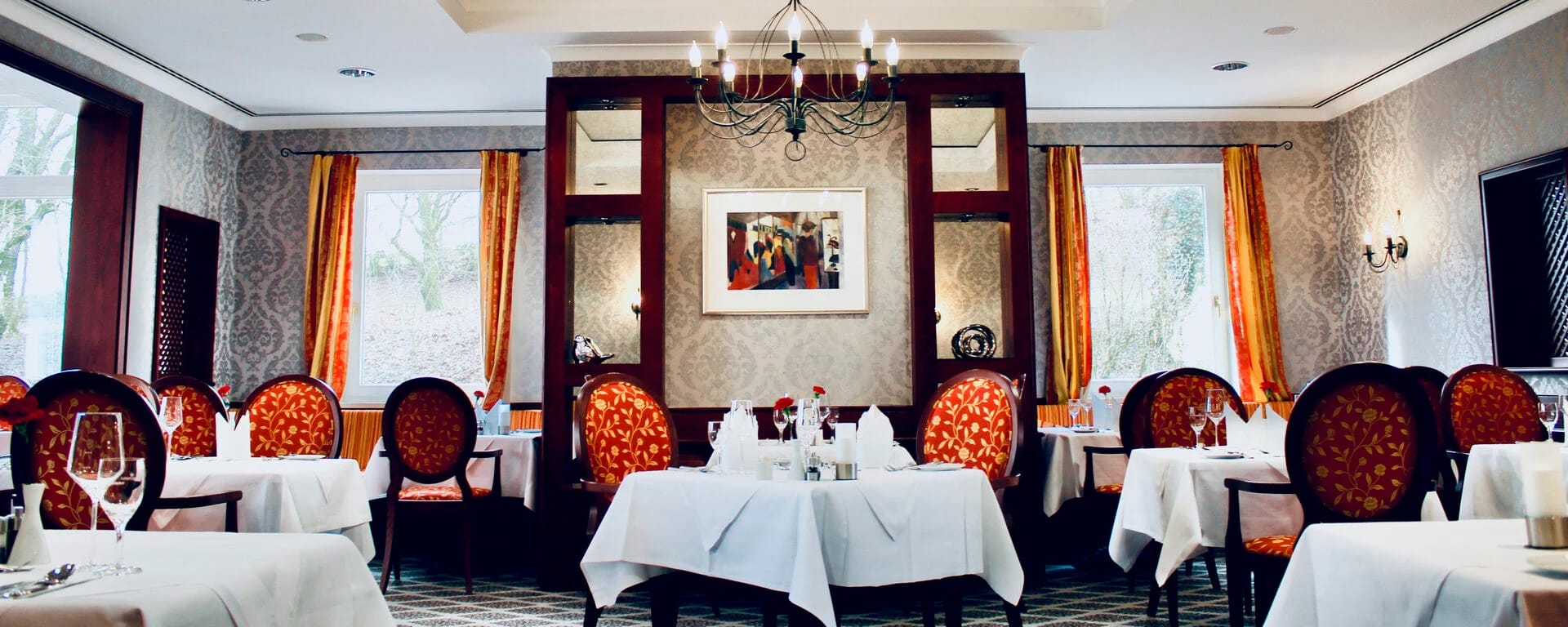 Restaurantstube mit weiß eingedeckten Tischen, roten Polsterstühlen und Kronleuchtern