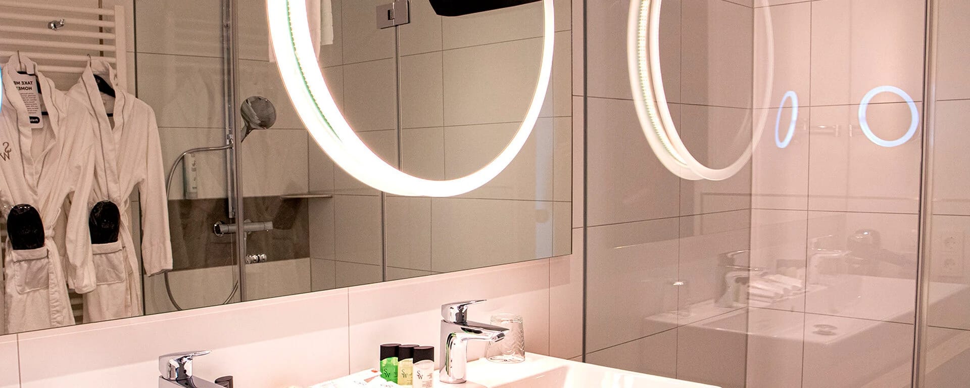 Badezimmer mit Doppelwaschtisch, runden Leuchtspiegeln und bereitliegenden Bademänteln