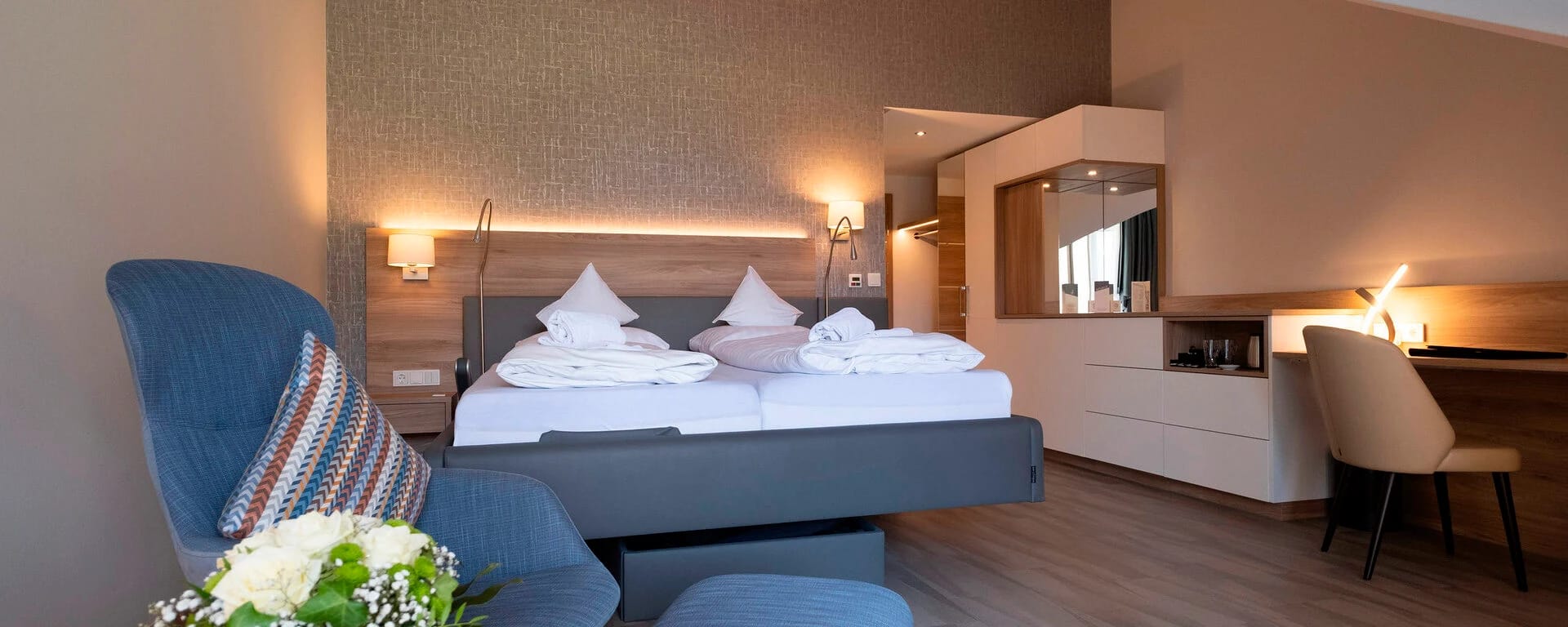 Doppelzimmer mit blauem Sessel, Schreibtisch und gemusterter Wand hinter dem Bett