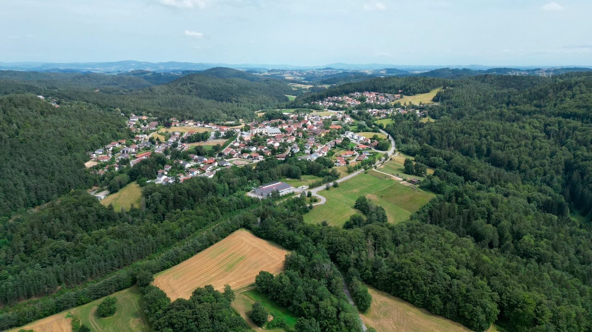 Luftaufnahme eines Dorfes zwischen bewaldeten Hügeln und Feldern