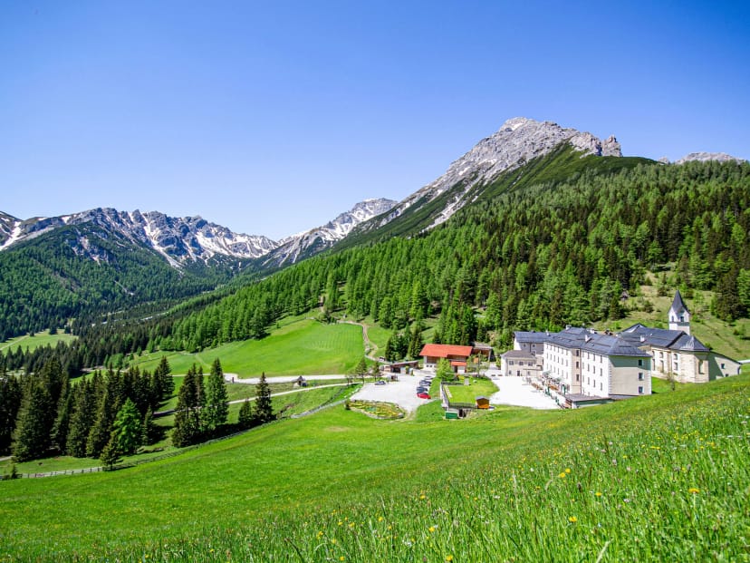 Klostergasthof und Klosterkirche auf einer Wiesenlichtung, umgeben von Nadelwald, dahinter Felsgipfel unter blauem Himmel