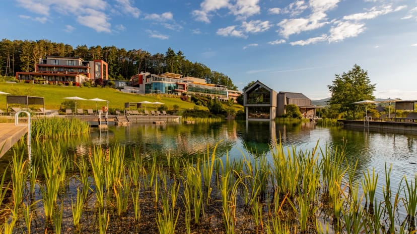 Resortgebäude am Naturbadeteich mit bewaldeten Hügeln bei Sonnenschein