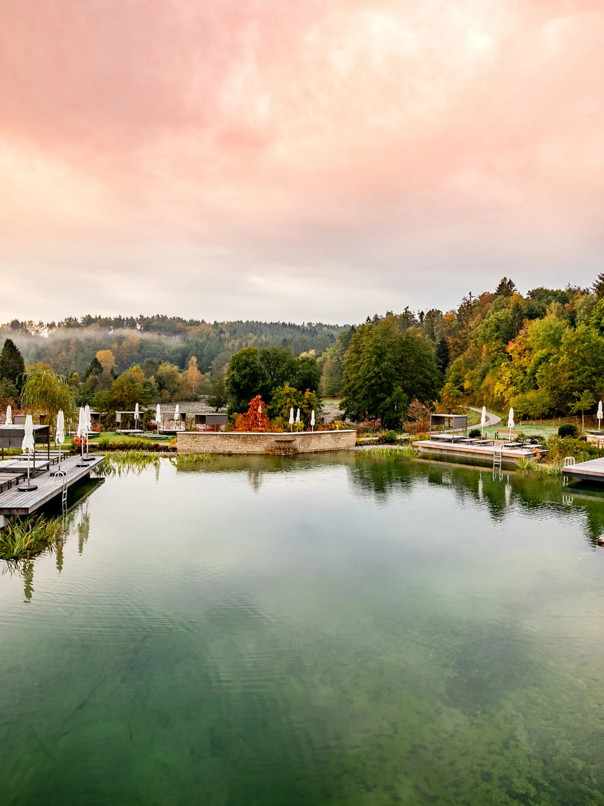 Naturbadeteich mit Liegeflächen und herbstlichem Ufer unter rosa Himmel