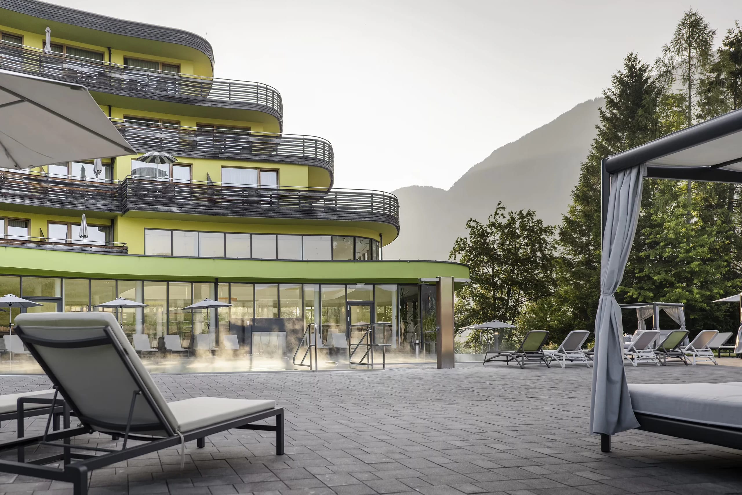 Geschwungene Fassade des Hotels DAS SIEBEN mit Balkonen, davor eine Poolterrasse mit Liegen und Daybed
