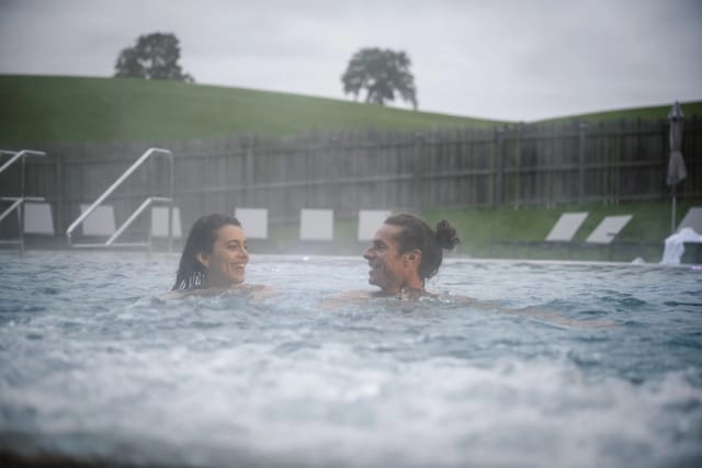 Außenansicht des geschwungenen grünen Hotelgebäudes mit Außenpool und Liegen vor Bergkulisse