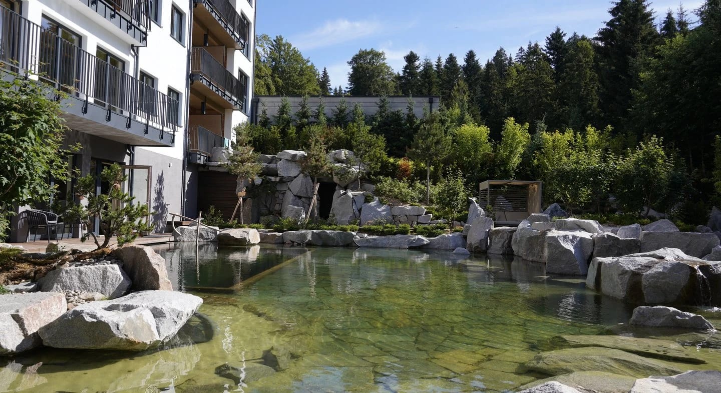 Naturbadeteich mit großen Felsen und Bepflanzung, links der Balkonflügel des Hauses