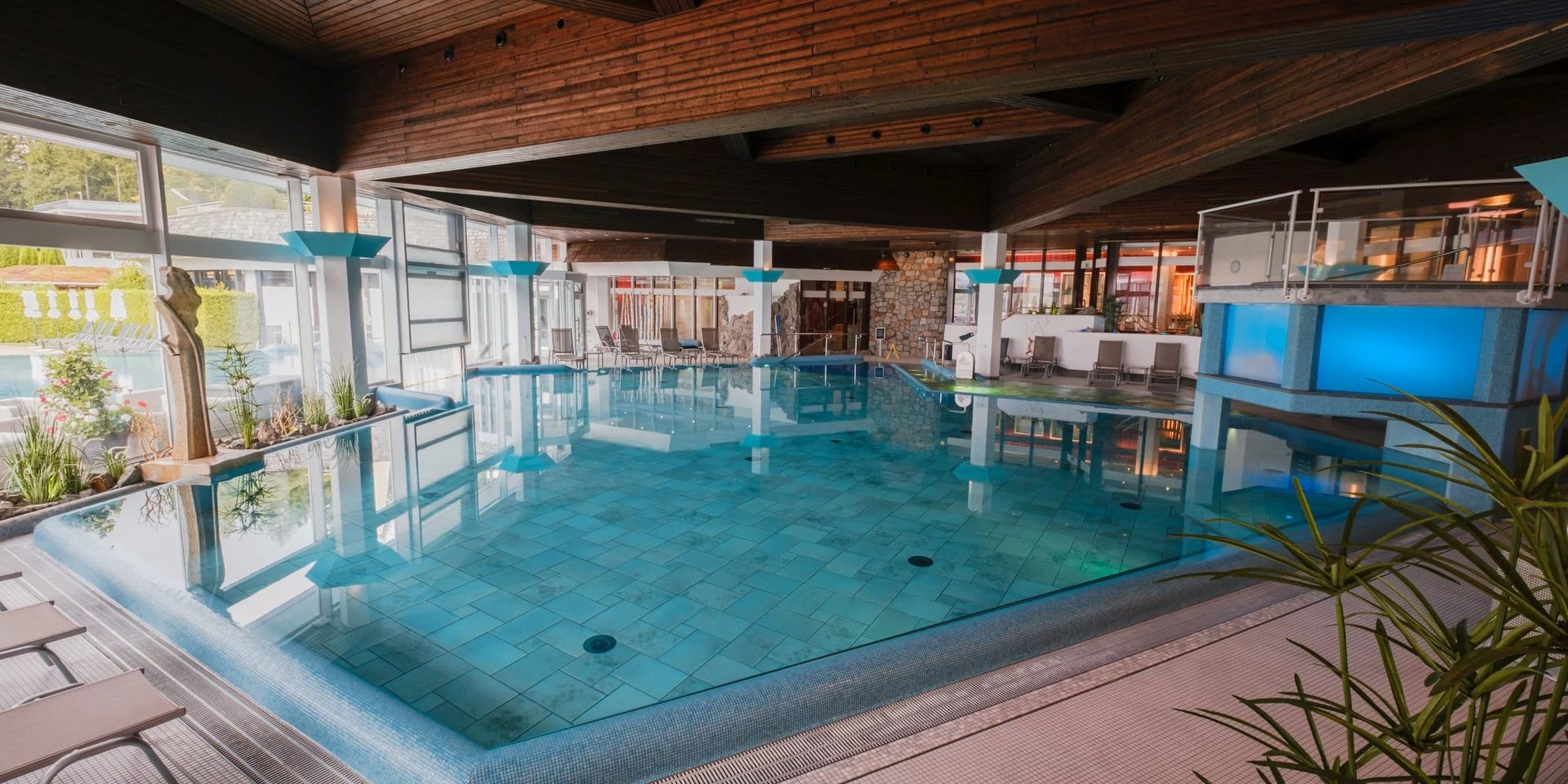 Hallenbad mit türkisfarbenem Becken, Holzdecke und breiter Fensterfront