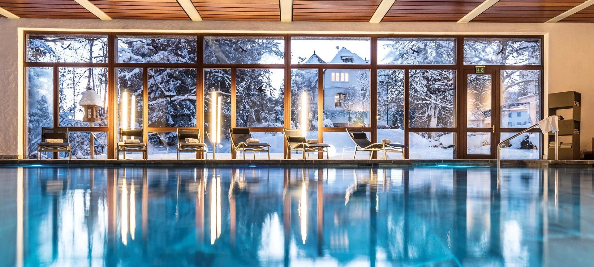 Hallenbad mit türkisem Wasser, Holzdecke, Liegen und Fensterfront zum verschneiten Wald