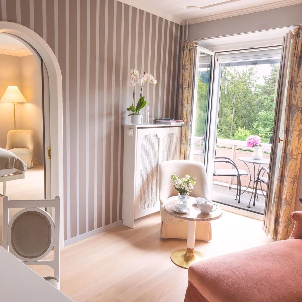 Zimmer mit gestreifter Wand, Bogenspiegel, Sessel und geöffneter Flügeltür zum Balkon
