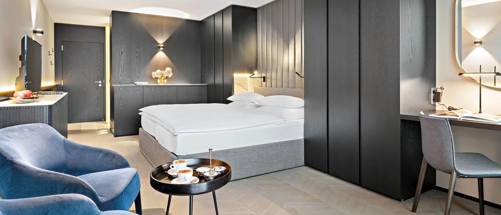 Modern eingerichtetes Zimmer mit Doppelbett, dunkler Schrankwand, blauem Sessel und Schreibtisch