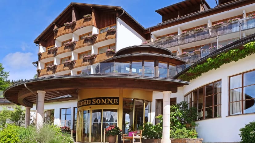 Vorderansicht des Hotels mit Holzbalkonen, Blumenkästen und überdachtem Eingang unter blauem Himmel