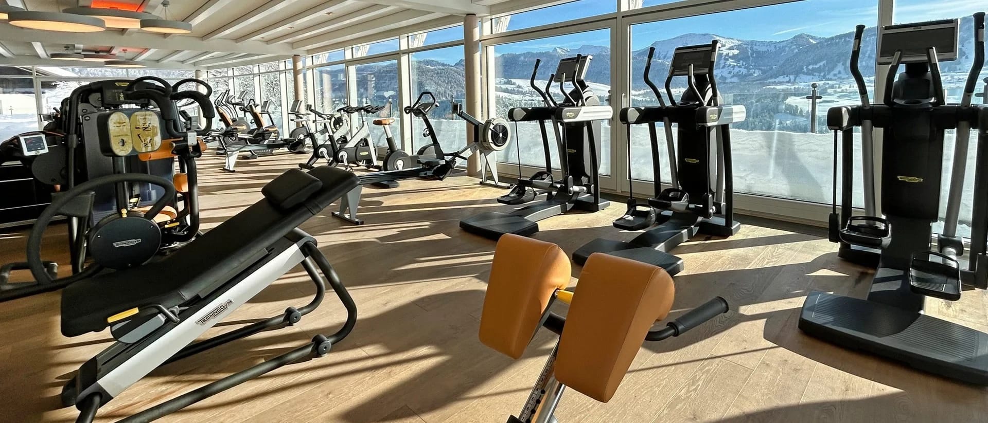Fitnessraum mit Ausdauergeräten und Panoramafenstern mit Blick auf schneebedeckte Berge