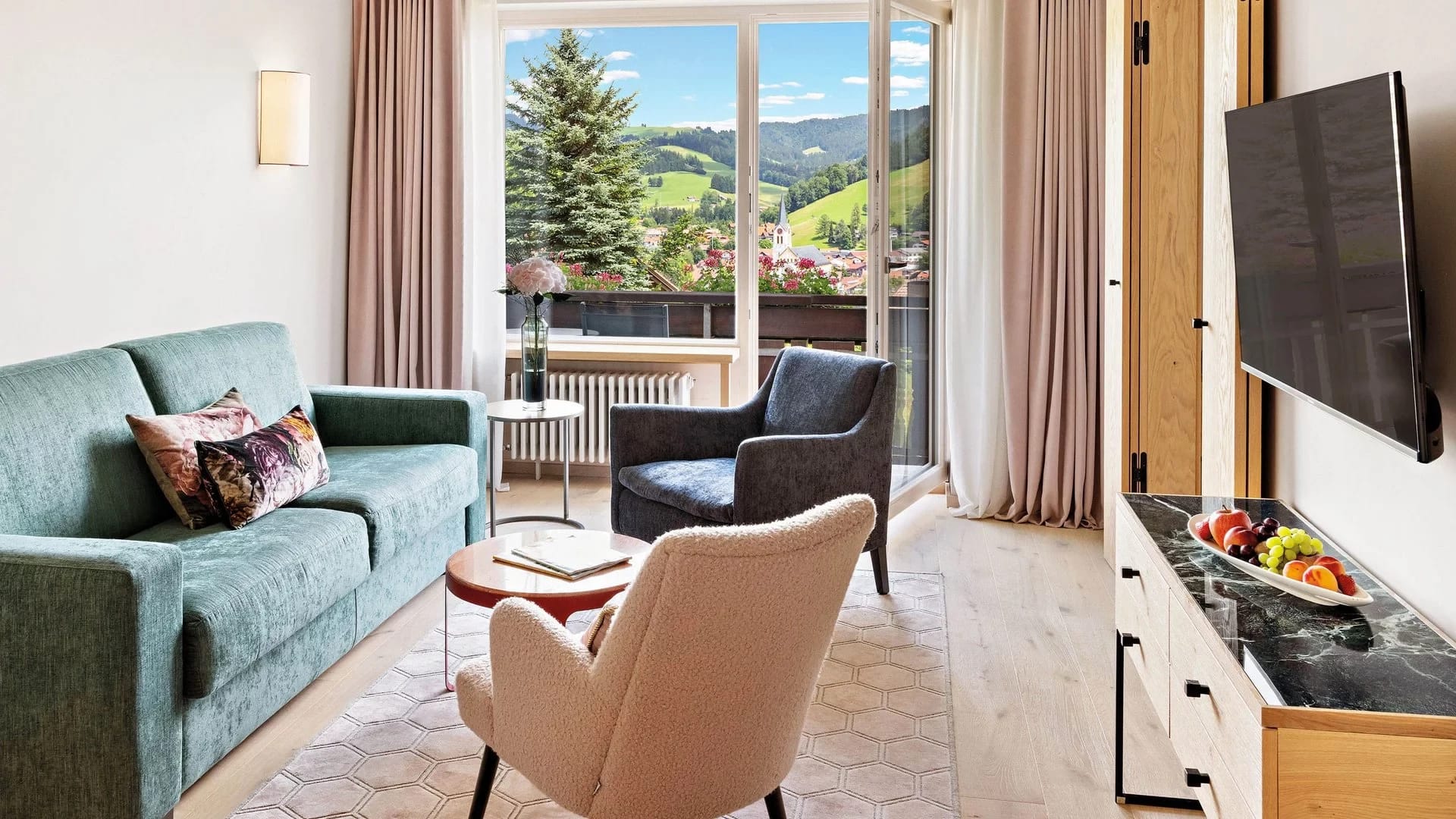 Wohnzimmer eines Appartements mit grünem Sofa, Sesseln und Balkon mit Blick auf Wiesen und Berge