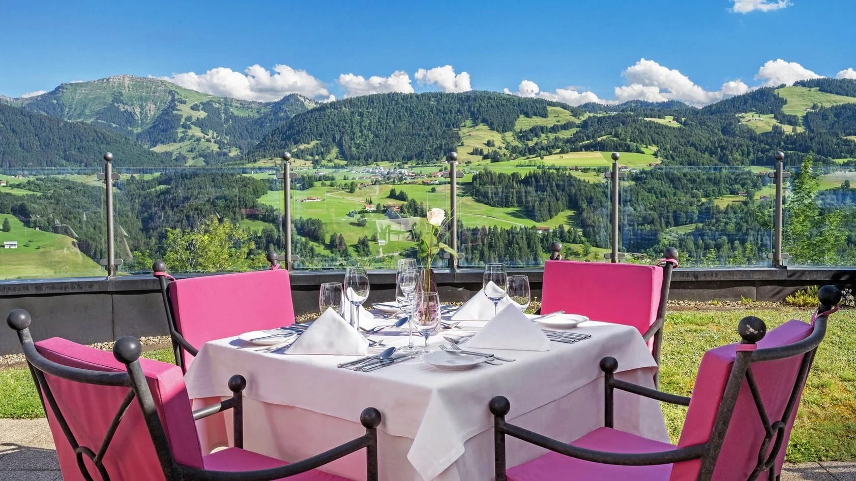 Gedeckter Tisch mit pinken Stühlen auf der Terrasse mit Blick über grüne Hügel und Berge