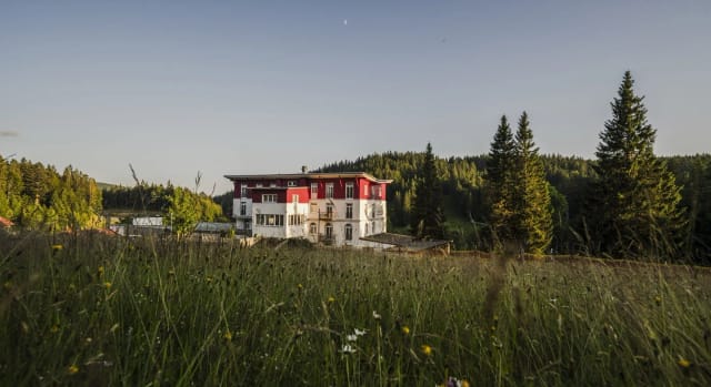 Das Hotelgebäude mit weiß-roter Fassade auf einer Wiese vor bewaldeten Hängen im Abendlicht