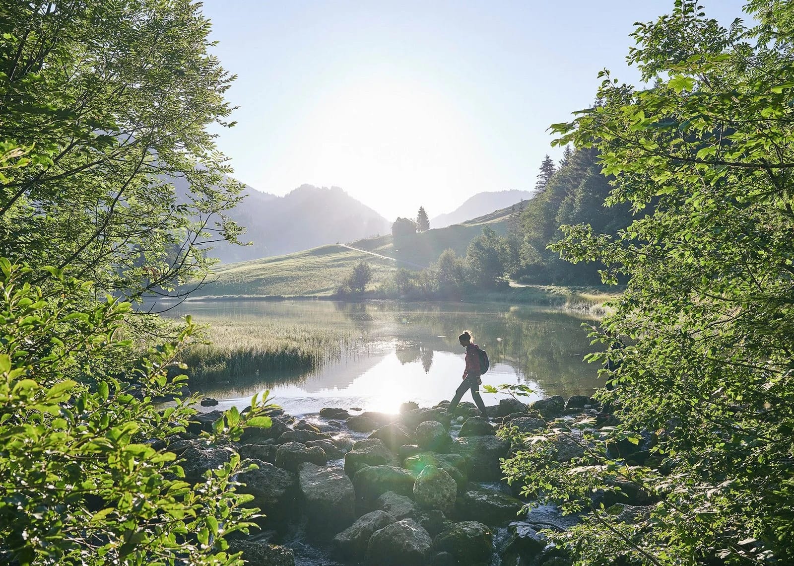 Wanderer am Ufer des Lecknersees, im Gegenlicht zwischen Bäumen, Wiesen und Bergen