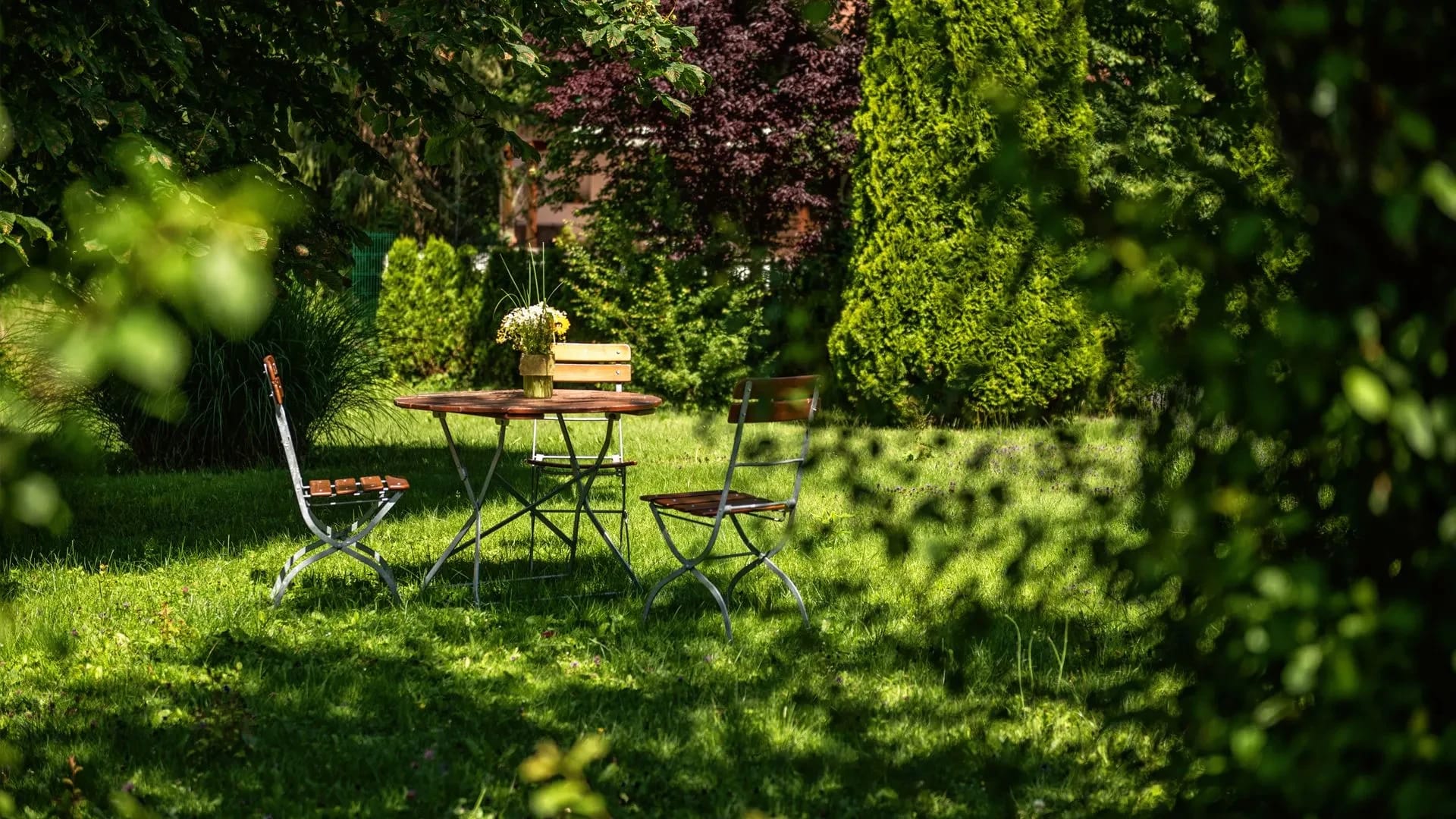 Sitzgruppe mit Tisch und zwei Stühlen auf der Gartenwiese zwischen Hecken und Bäumen