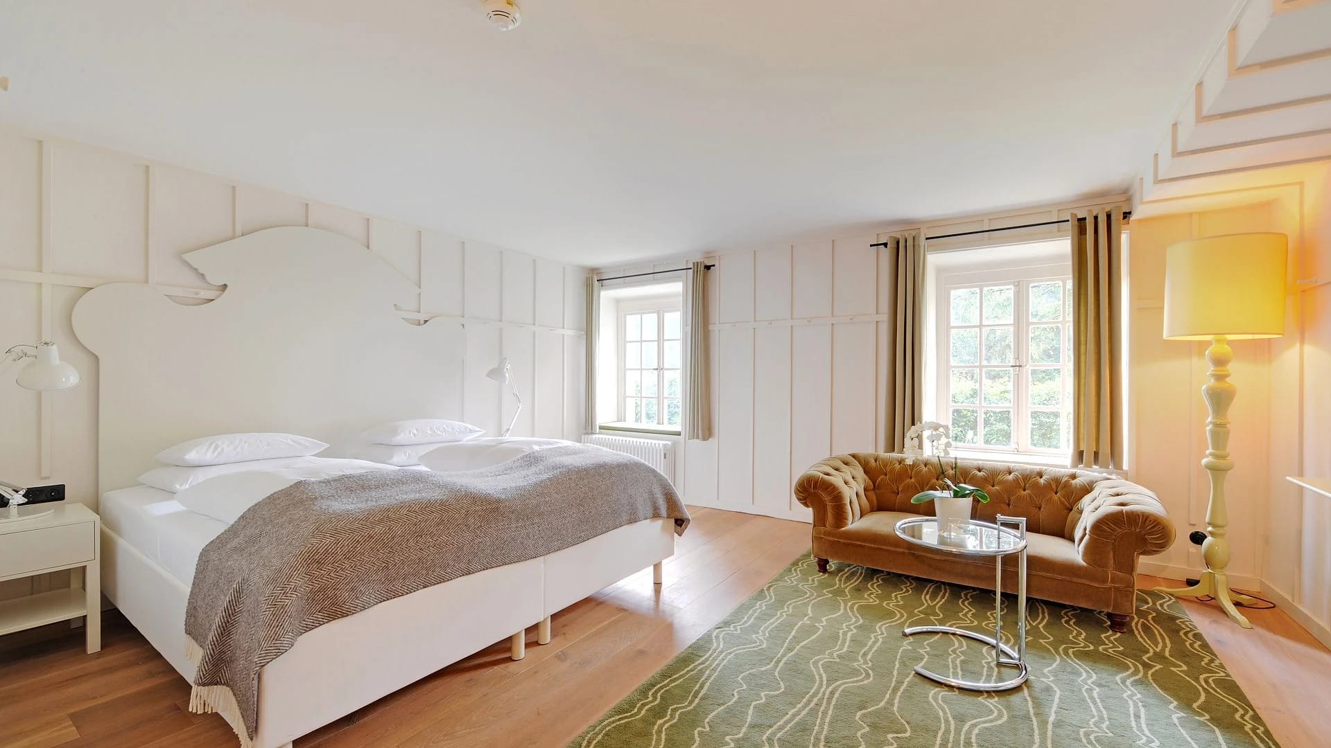 Weiß getäfeltes Zimmer mit Doppelbett, cognacfarbenem Ledersofa und gemustertem Teppich
