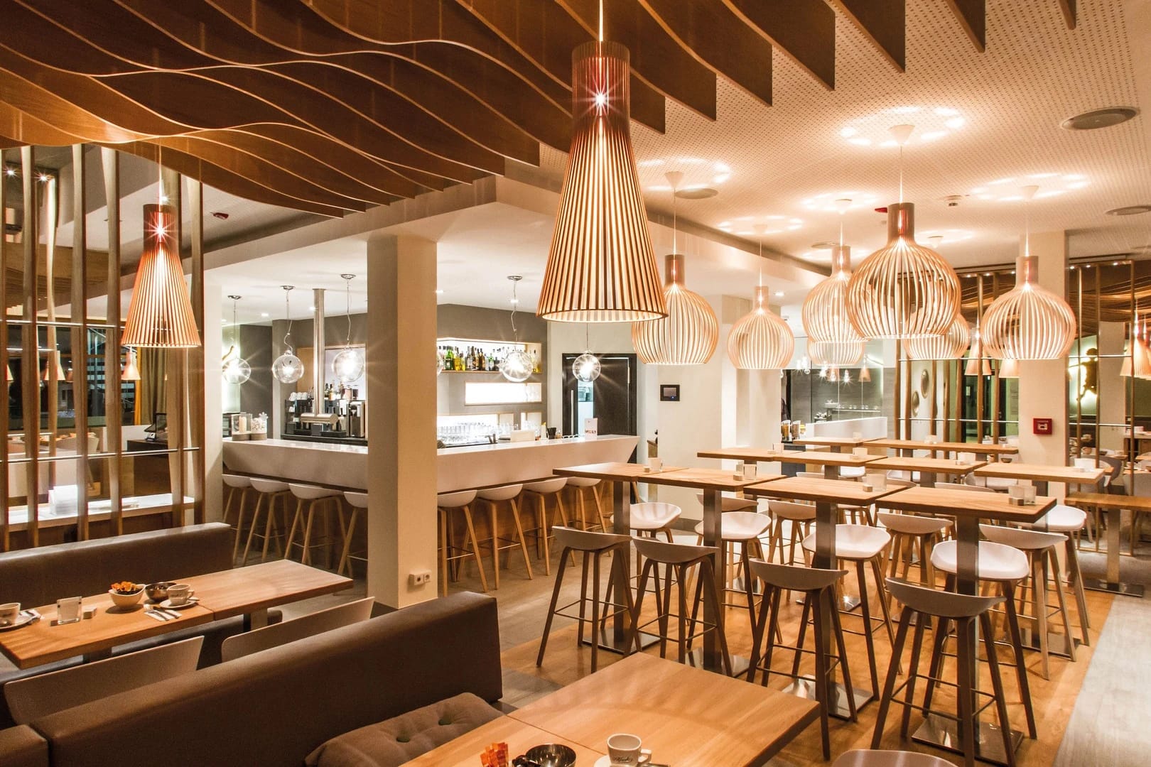 Restaurant mit geschwungener Holzdecke, Pendelleuchten, Tischen und Bartresen
