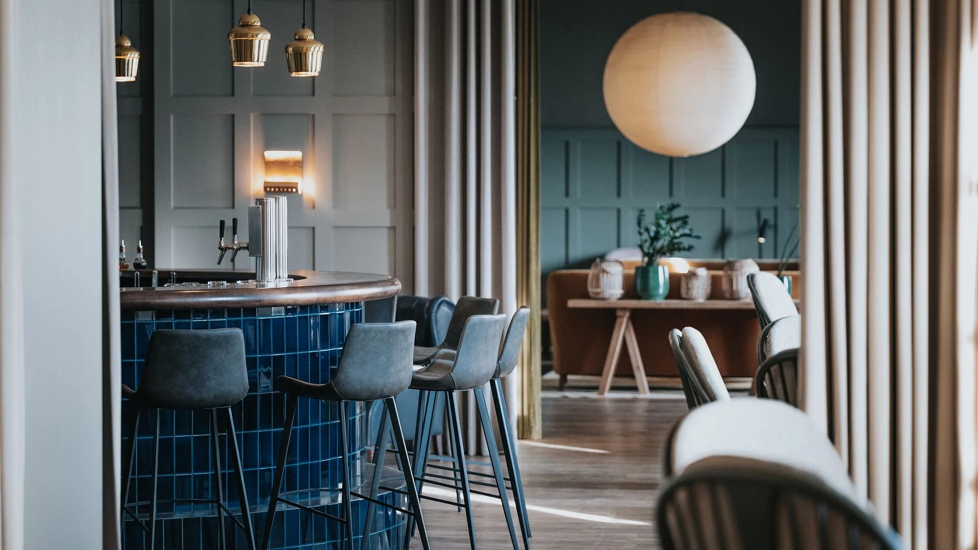 Bar mit blau gefliester Theke, Barhockern und Messingleuchten, dahinter ein Sitzbereich