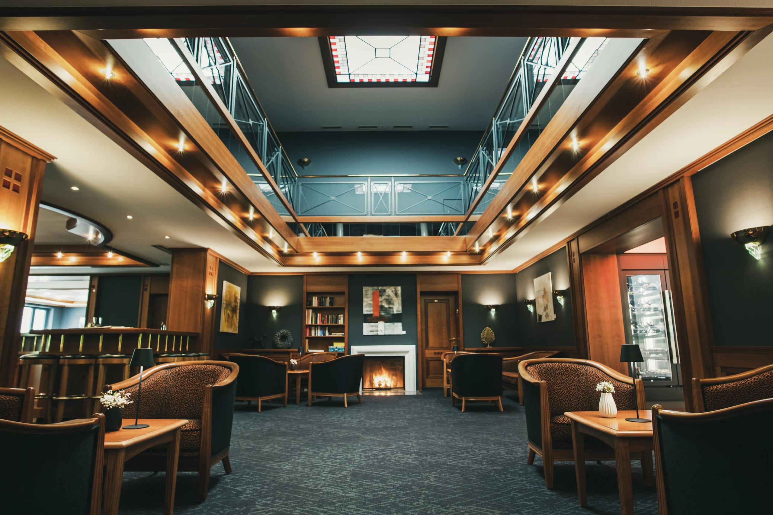 Hotellobby mit Kamin, Sesseln, Holzvertäfelung und umlaufender Galerie unter einem Glasdach