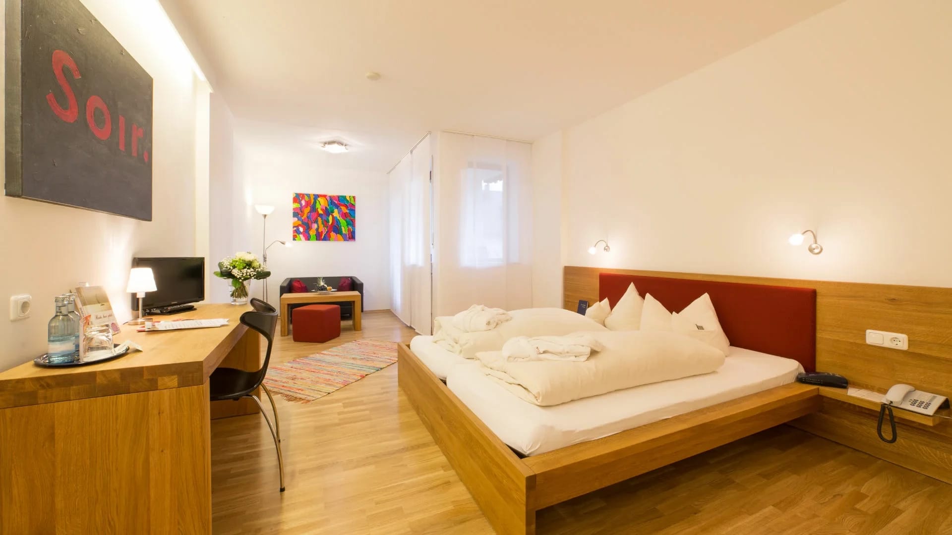 Modernes Doppelzimmer mit Holzboden, Schreibtisch, Sitzecke und Bett mit rotem Kopfteil
