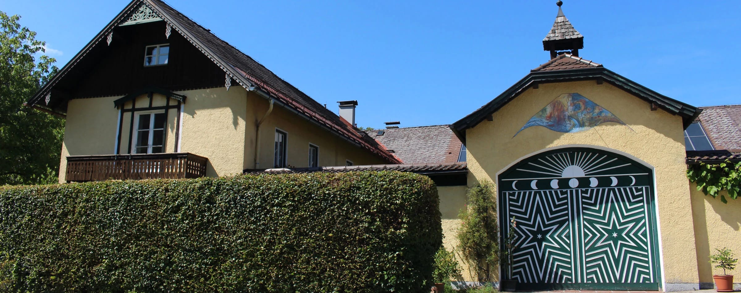 Eingangstor mit Sternenmotiv und Türmchen neben dem Wohngebäude mit Balkon, davor eine hohe Hecke