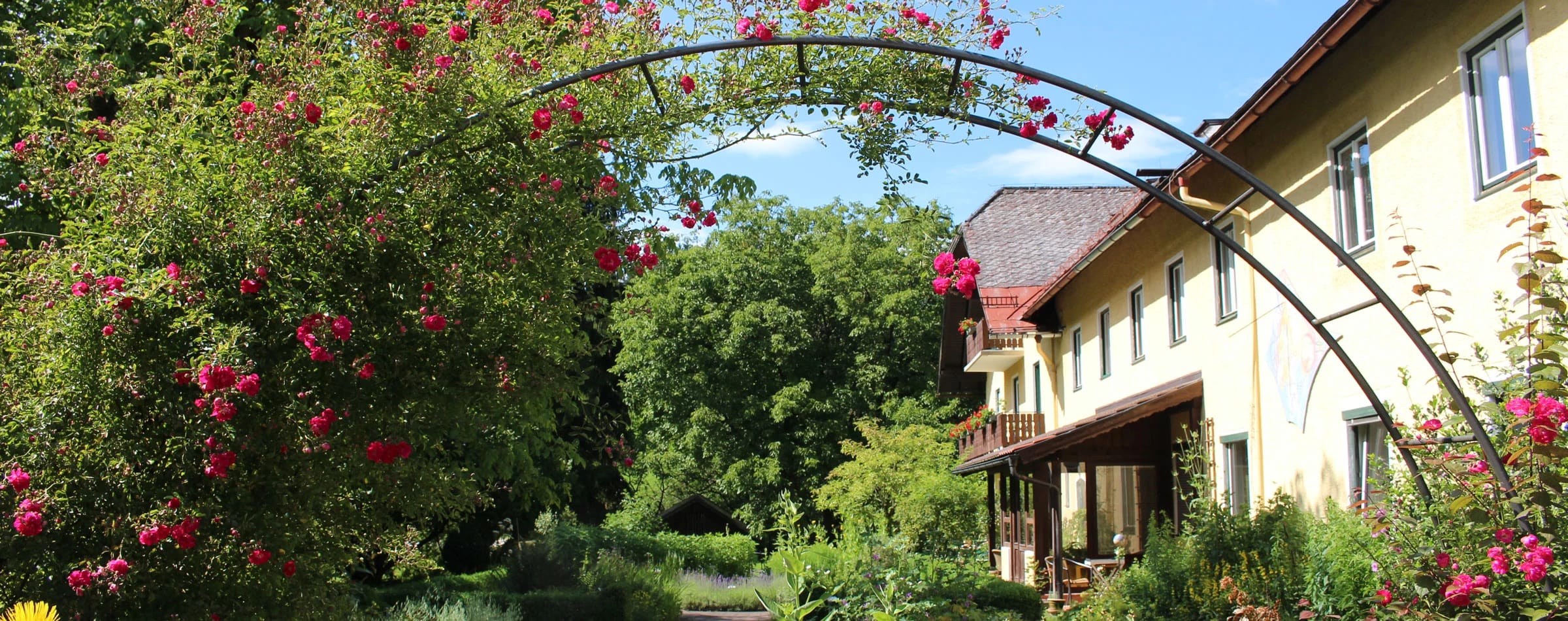 Rosenbogen im Garten mit Blick auf die lange Hausfassade mit Holzbalkon