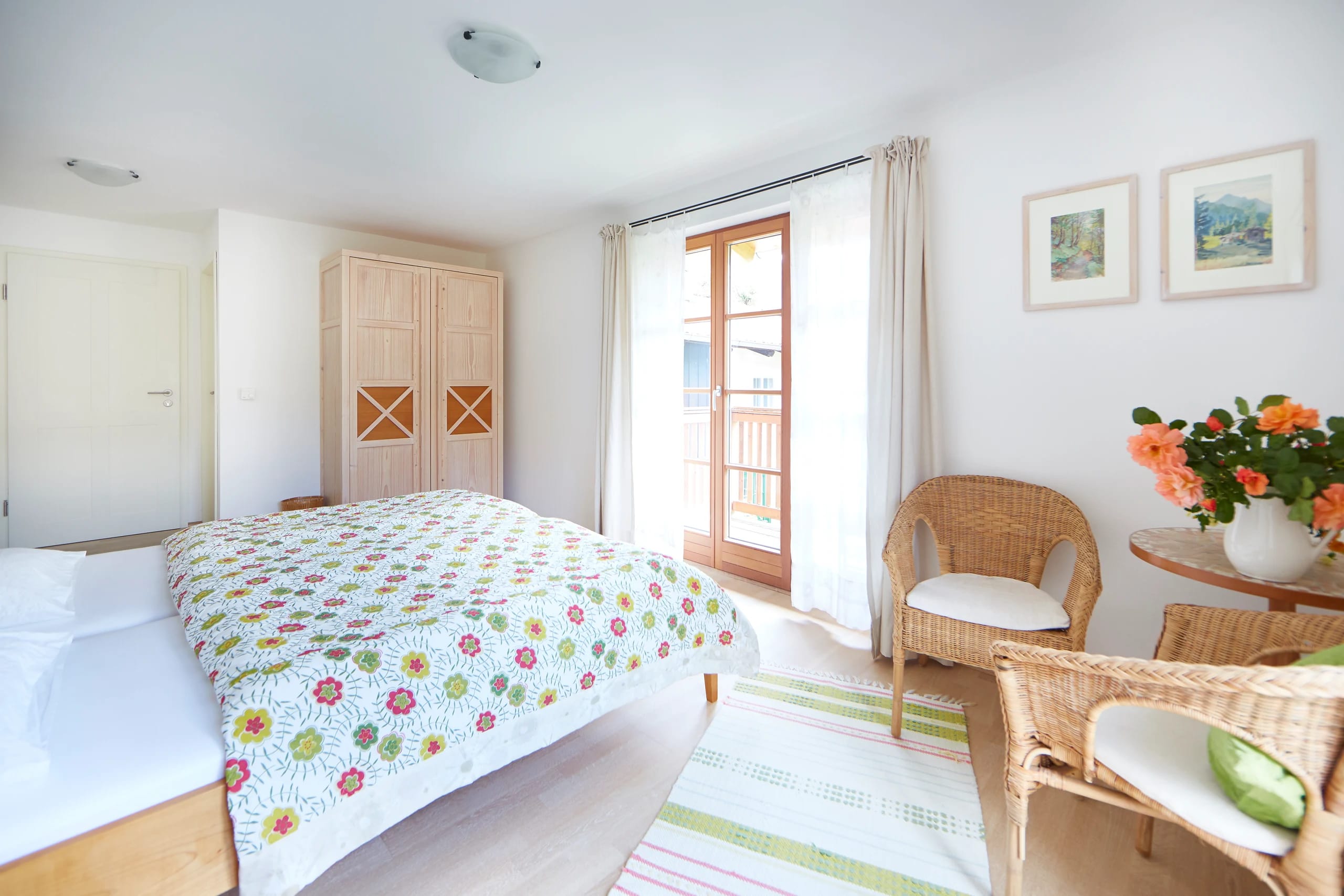 Doppelzimmer mit geblümter Tagesdecke, Holzschrank, Korbsesseln und Balkontür