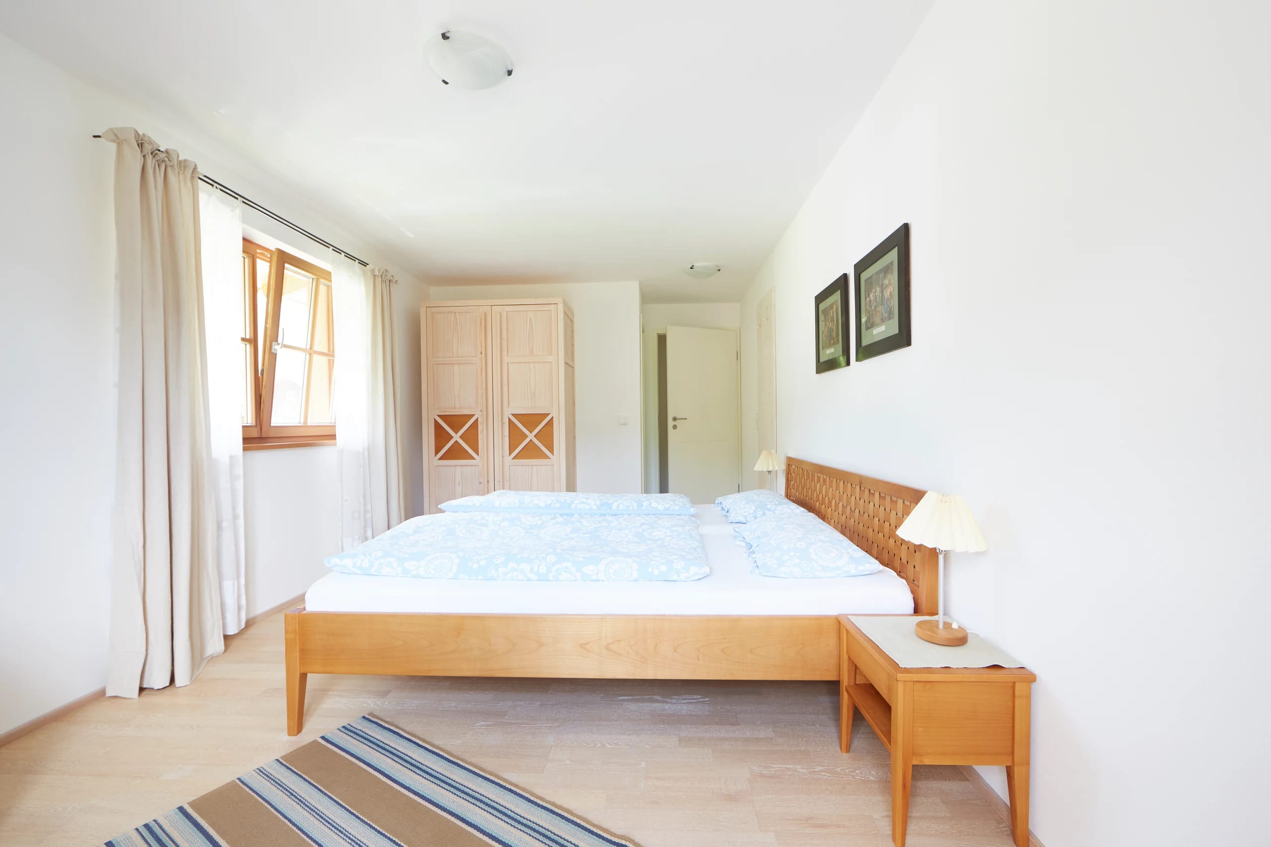 Doppelzimmer mit hellblauer Bettwäsche, Holzschrank und Fenster mit langen Vorhängen