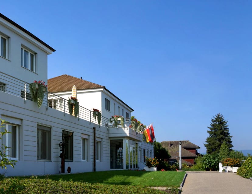 Weiss verputztes Haupthaus mit Balkonen, Blumenkästen und Fahnen, davor Rasenfläche und Gartenweg