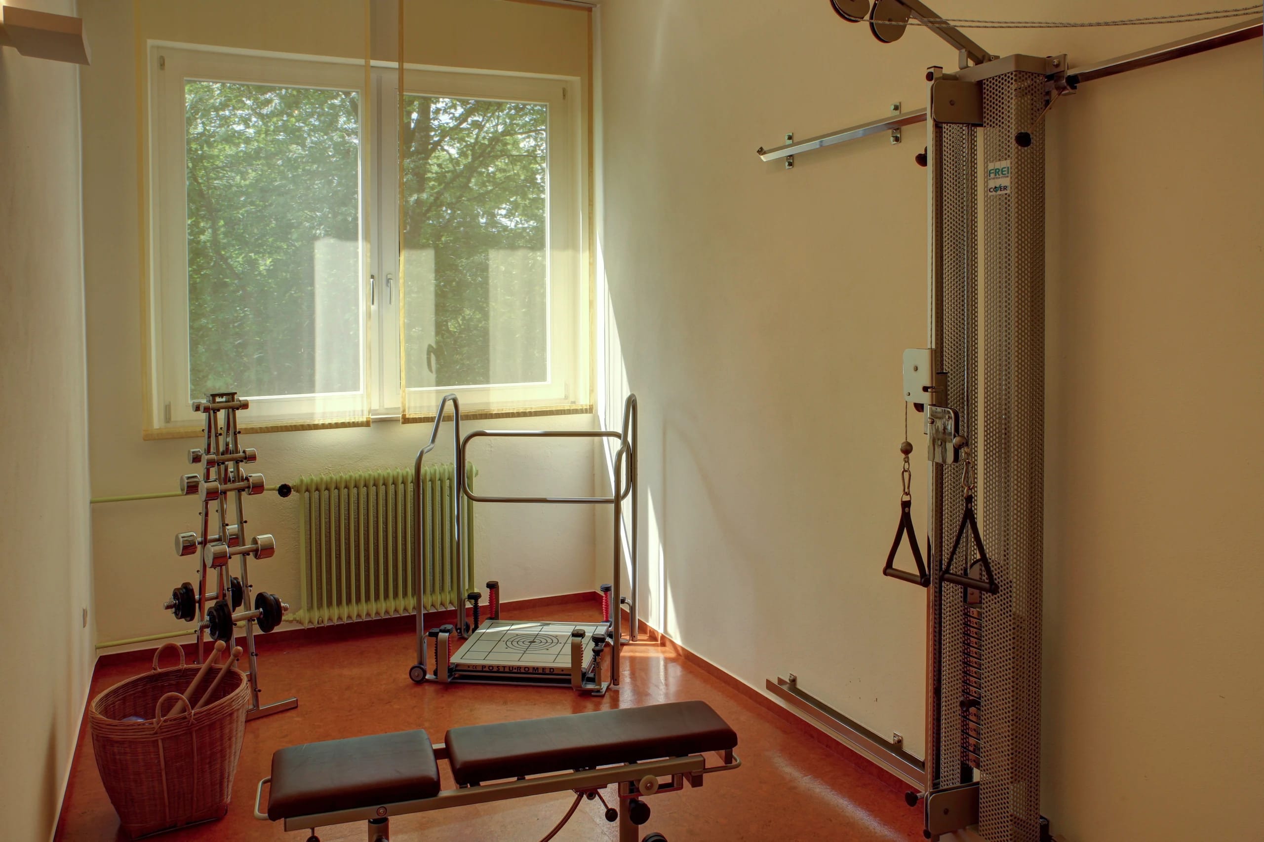 Kleiner Fitnessraum mit Zugapparat, Hantelbank und Gewichten am Fenster