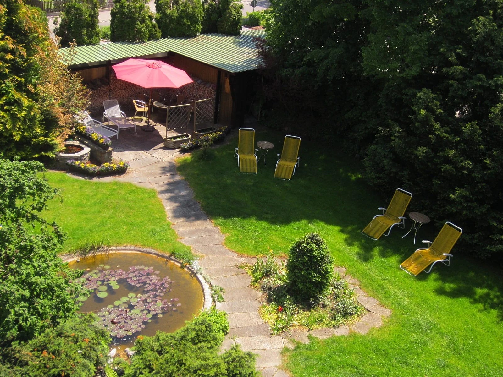 Blick von oben auf den Garten mit Rasen, kleinem Teich, überdachter Sitzecke mit rotem Sonnenschirm und gelben Liegestühlen