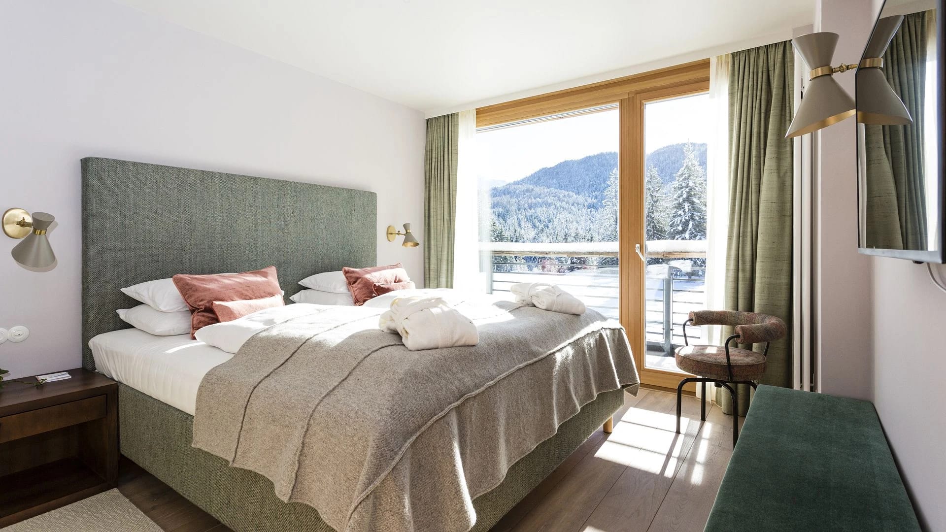 Schlafzimmer mit grün gepolstertem Bettkopfteil und bodentiefem Fenster mit Blick auf verschneite Berge