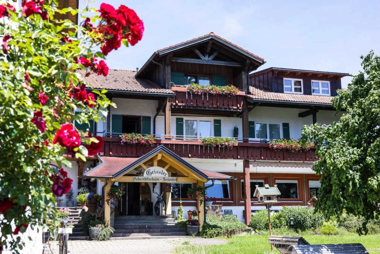 Haupthaus mit Holzbalkonen, Blumenschmuck und überdachtem Eingang, im Vordergrund rote Rosen