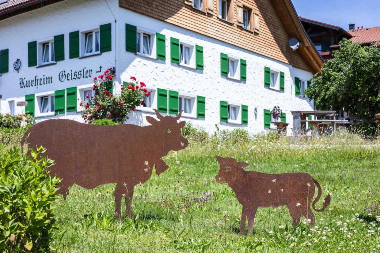 Weiß getünchtes Bauernhaus mit grünen Fensterläden und Blumenkästen, davor eine Wiese mit Rinder-Silhouetten aus Metall