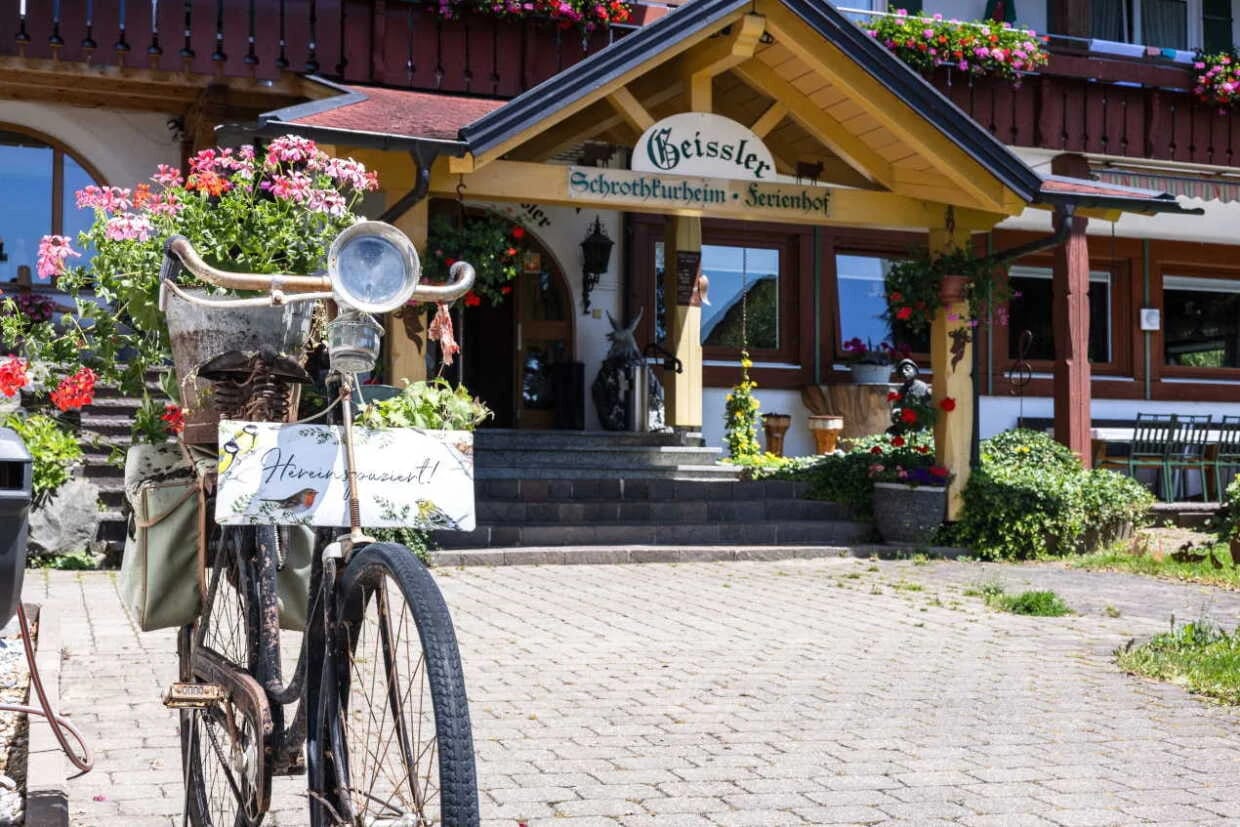 Hauseingang mit Holzvordach und Schriftzug, davor ein altes Fahrrad mit Blumenschmuck