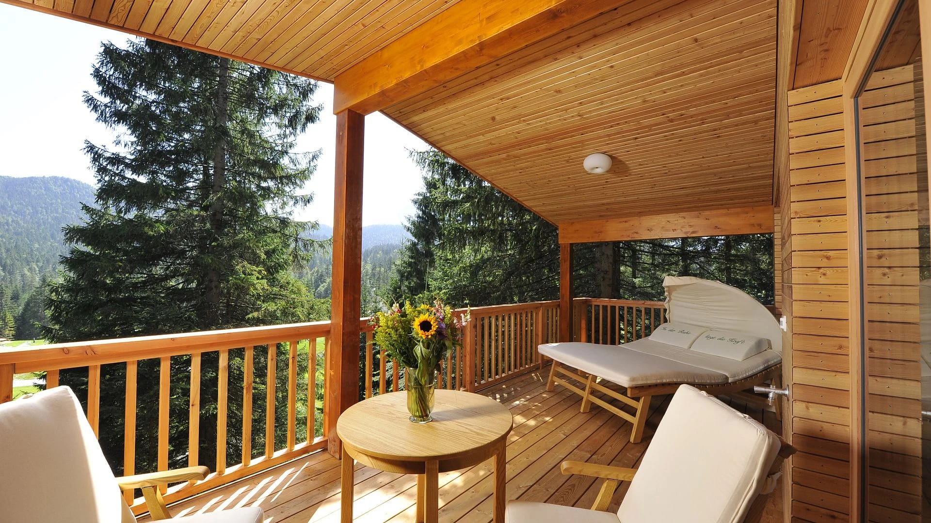 Überdachte Holzterrasse mit Doppelliege und kleinem Tisch, Blick über Nadelwald zu den Bergen