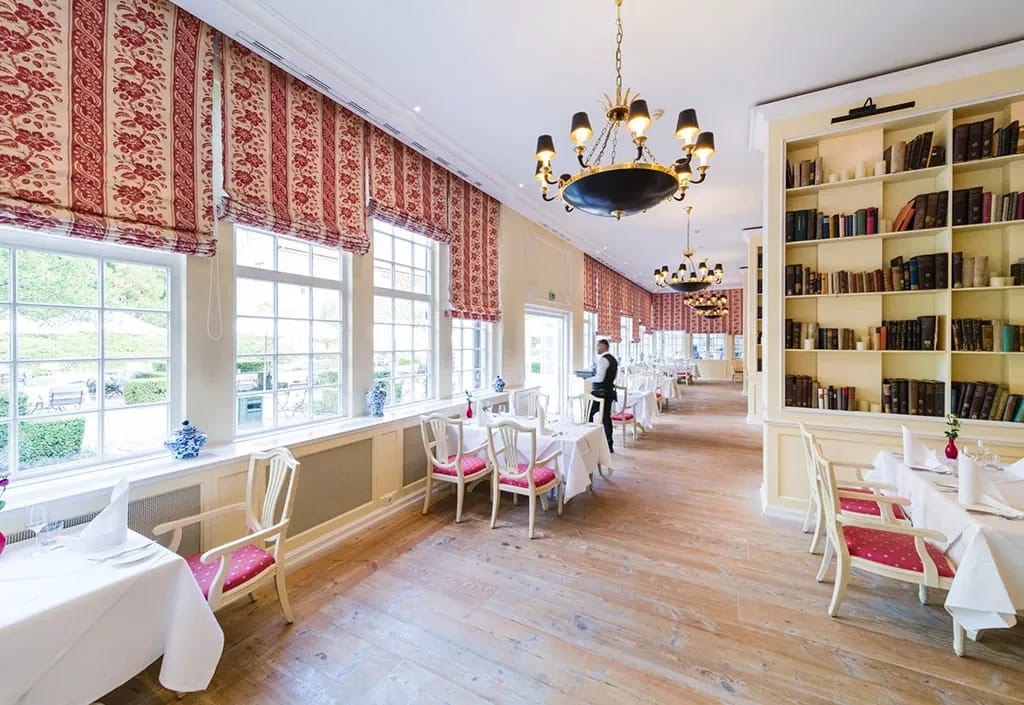 Restaurantsaal mit eingedeckten Tischen, Bücherregalen und rot gemusterten Vorhängen