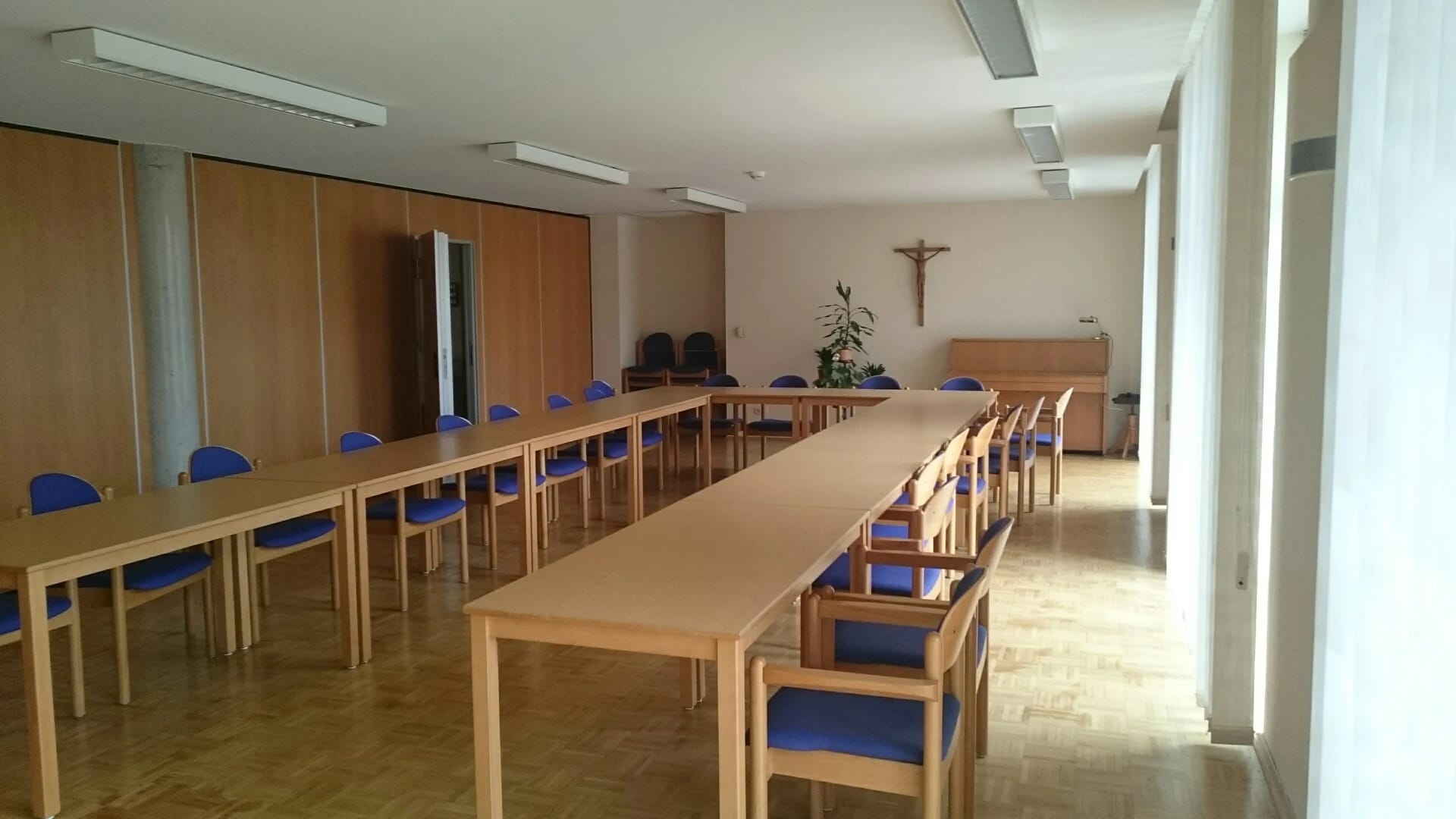 Heller Saal mit langen Holztischen, blauen Stühlen und Kreuz an der Stirnwand
