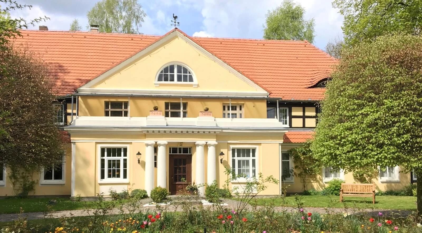 Gelbes Gutshaus mit rotem Ziegeldach und Säulenportal, davor ein bepflanztes Beet