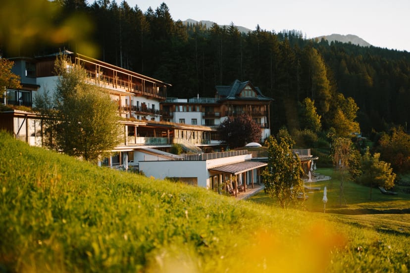 Das Hotel im Abendlicht, eingebettet in eine grüne Wiese vor bewaldeten Bergen