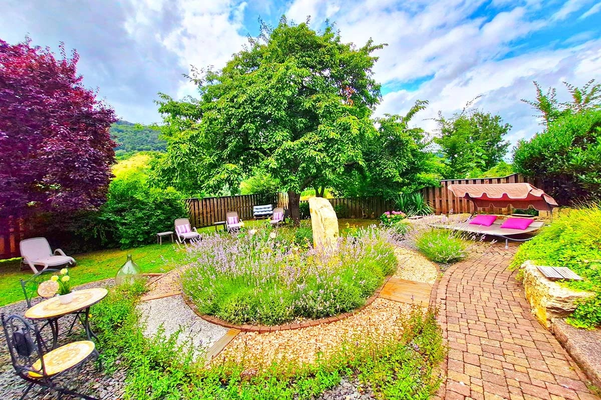 Gartenanlage mit Liegen, Sitzplätzen und Lavendelbeet unter einem großen Baum
