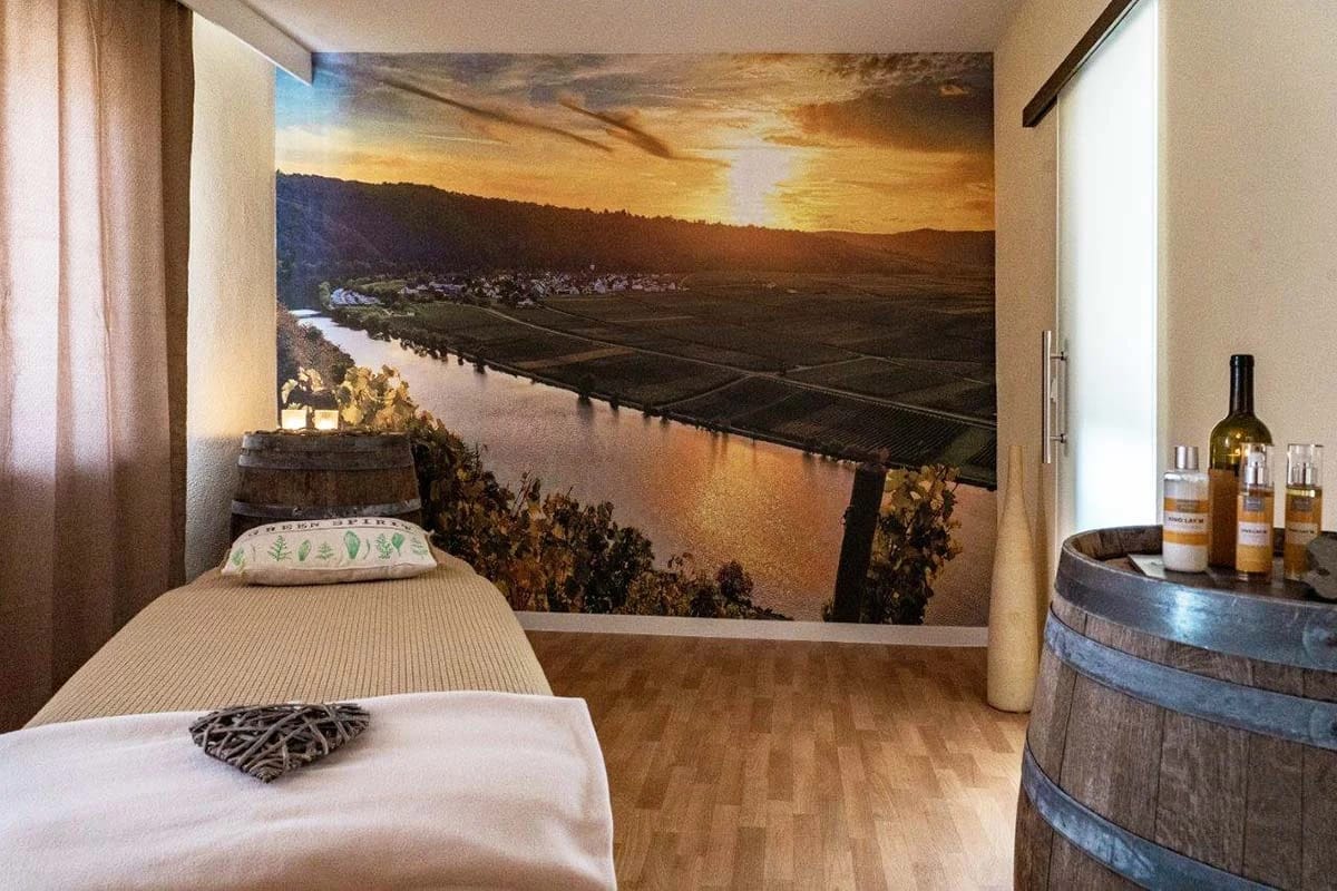 Behandlungsraum mit Massageliege, großem Wandbild vom Moseltal und einem Weinfass mit Flaschen darauf