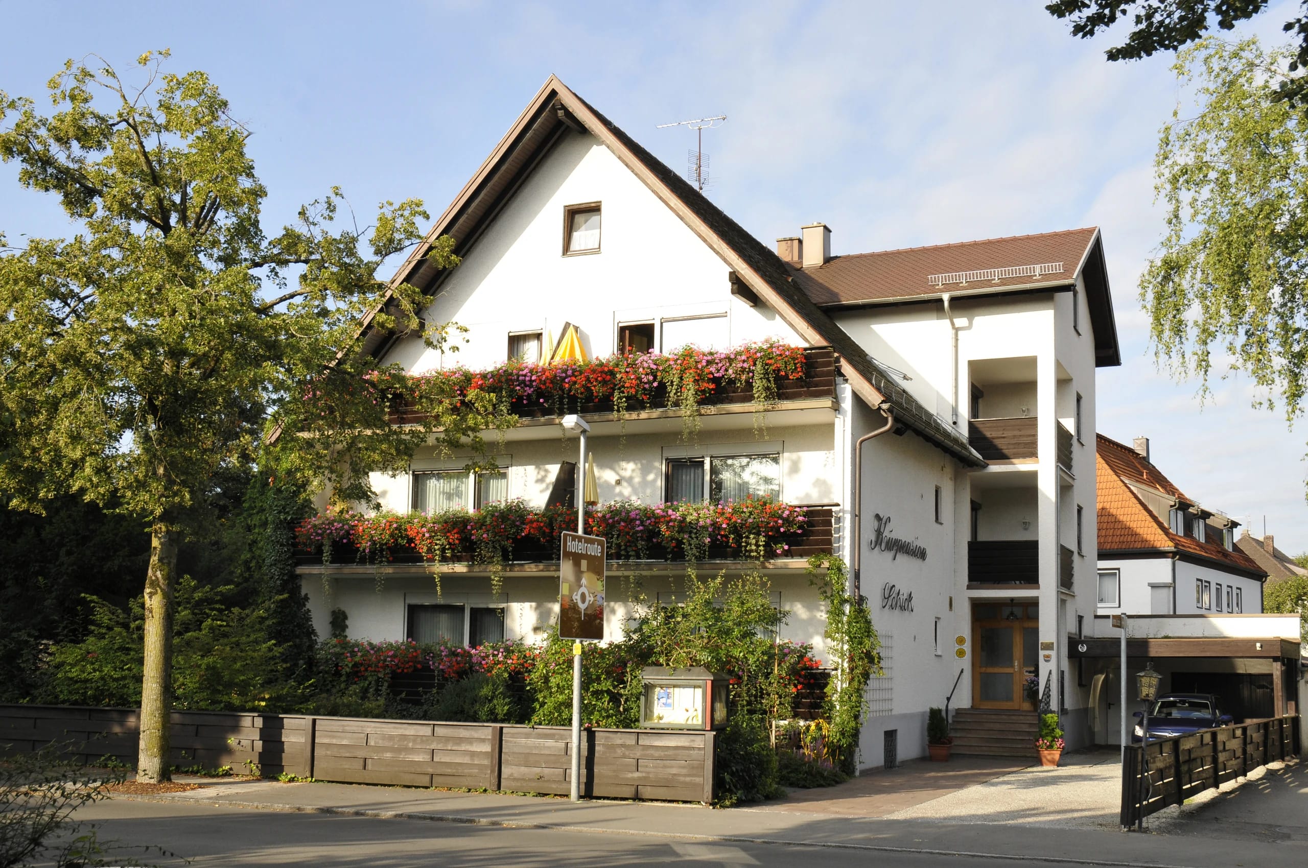 Weiß verputztes Haus mit spitzem Giebel, Balkonen voller roter und orangefarbener Blumen, Bäumen davor und dem Hauseingang